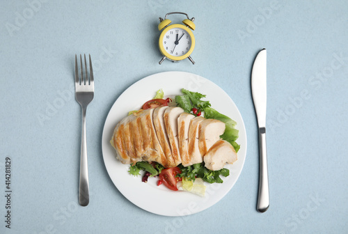 Plate of appetizing food, alarm clock and cutlery on light blue table, flat lay. Nutrition regime