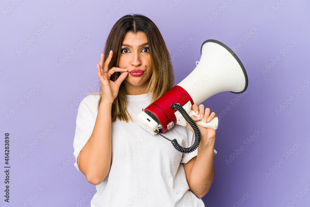 Young indian woman holding a megaphone isolated with fingers on lips keeping a secret.