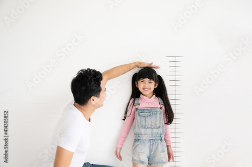 Dad measures height increase of her child daughter at white brick wall with scale