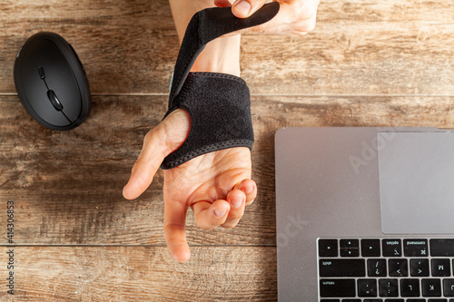 Chronic trauma to the wrist joint  in people using computer mouse may lead to disorders that cause inflammation and pain. A woman working on desk uses wrist support brace and ergonomic vertical mouse photo