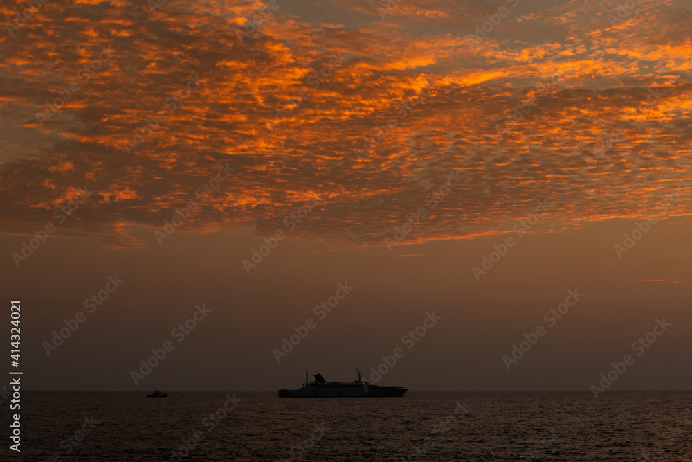 silhouetted of a long distanced ship in horizon of the ocean, after the sunset golden hour photograph, clouds glowing in the evening sunlight.