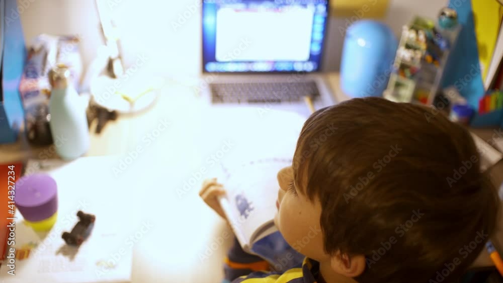 Back view of caucasian six year's old boy, studying at his desk with distance education system during coronavirus lockdown. High view over the shoulder