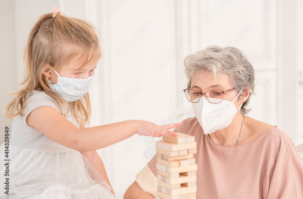 Happy family during quarantine Coronavirus (Covid-19) epidemic. Senior grandmother wearing protective face mask plays with her little granddaughter