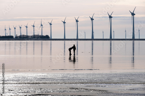 Silhouette of two unrecognizable boys playing on and reflected in the ice of the frozen bay of Lemmer, the Netherlands. In the background windmills for generating energy 