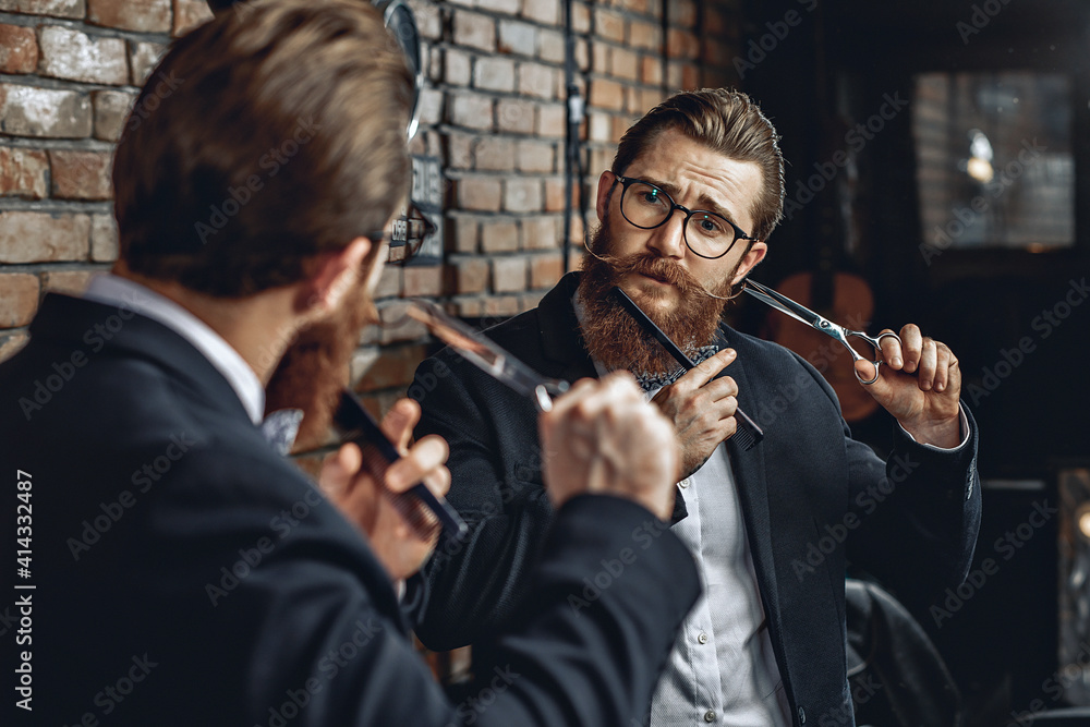 Attractive charming male barber in glasses, with a mustache and a beard, looking in the mirror and holding a comb and scissors next to his face. Barbershop concept
