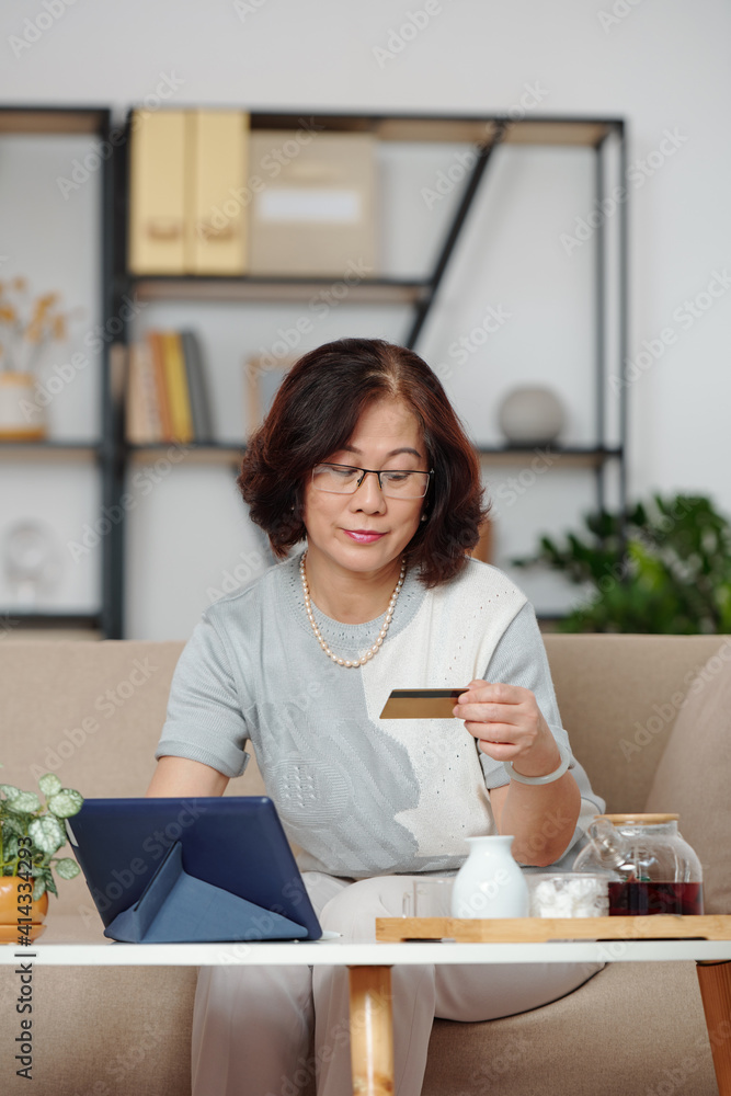 Asian senior woman entering information from credit card when drinking tea and shopping online via tablet computer Asian senior woman entering information from credit card when drinking tea and shopping online via tablet computer