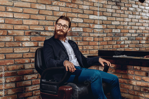 Stylish man in a jacket, glasses, with a mustache and a beard, sitting on a leather armchair and looking away. Barber shop. Style concept