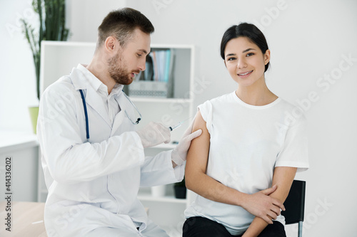 the doctor injects the covid-19 vaccine into the shoulder of the patient in a white t-shirt on a light background