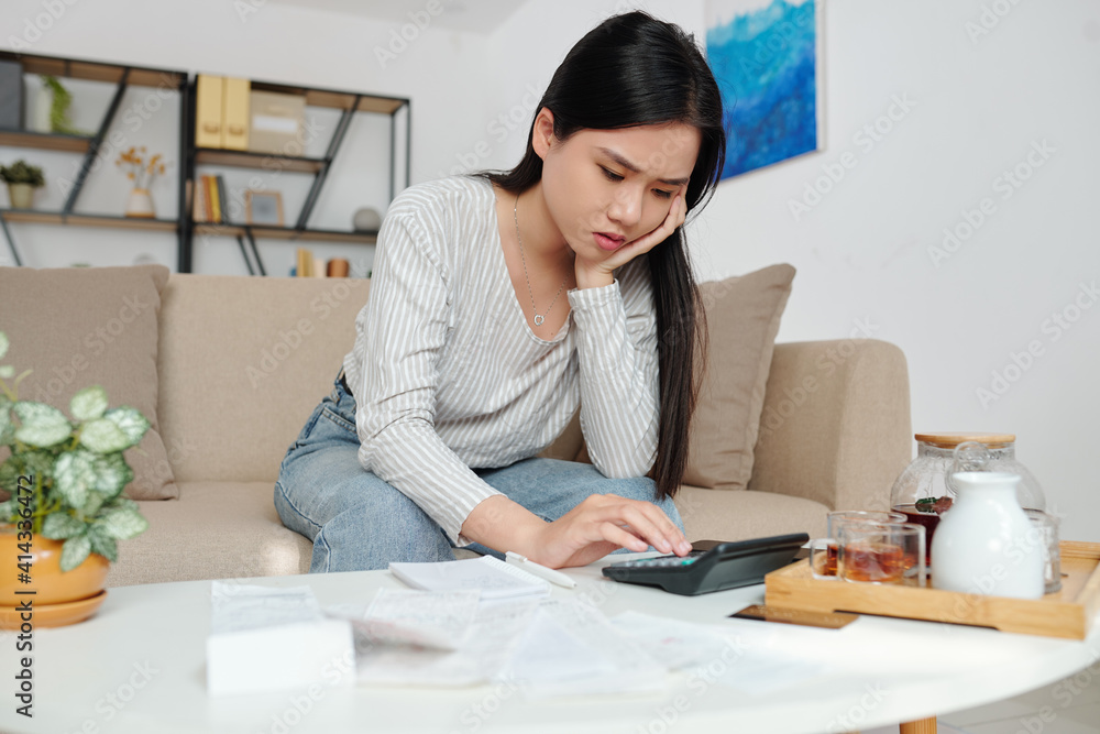 Stressed unhappy young Vietnamese woman calculating her monthly taxes, bank account balance and credit card payment bills