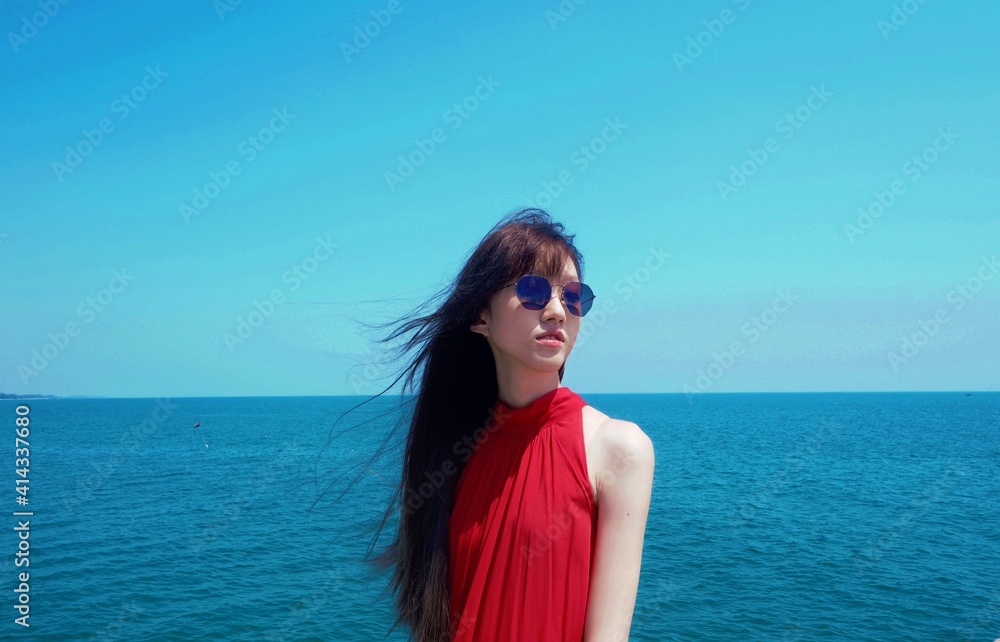 Asian girl in red dress wearing sunglasses looking up the sky while the sea breeze blows her hair.