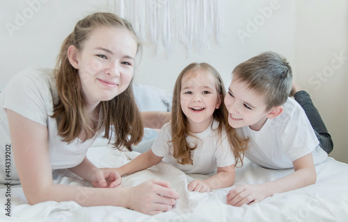 The concept is happy brother and sister. Three children of different ages: a teenage girl, a little girl and a boy. Children in white T-shirts lie on the white bed and smile.