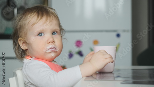 Side view little cute white girl blonde with pink bib drinks kefir from pale pink glass and looks at camera. Mouth in kefir or yogurt. Baby wear white shirt or bodysuit.