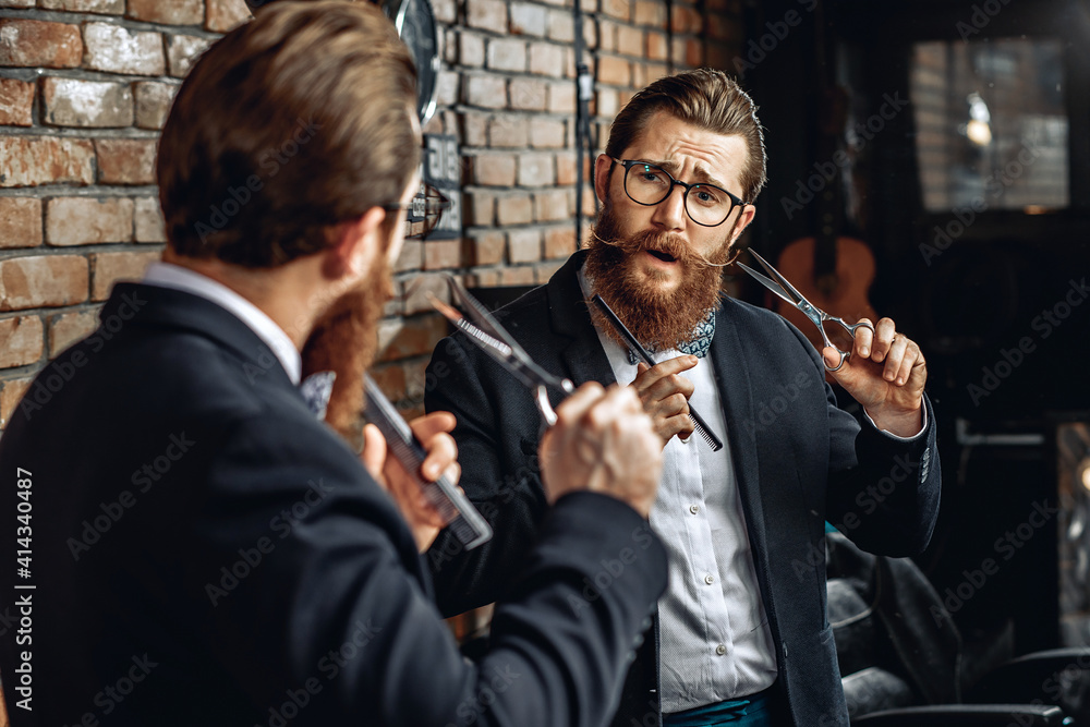 Portrait of a handsome stylish barber with glasses holding comb and scissors and looking in mirror holding tools next to beard and mustache. Luxury concept
