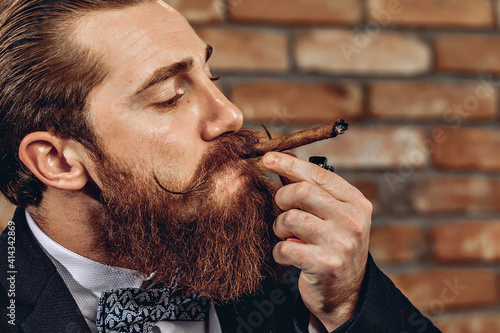 Cuadro en lienzo Close up portrait of attractive pretentious man with a mustache and beard smoking a brown cigar against a brick wall background