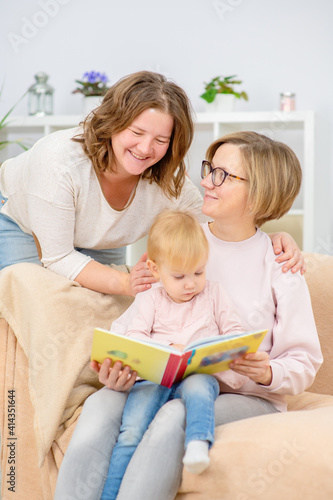 Two young women are sitting in the living room on the couch with a child on their knees and are reading a book to the child. Gay lesbian couple at home with a baby. Lesbian family concept