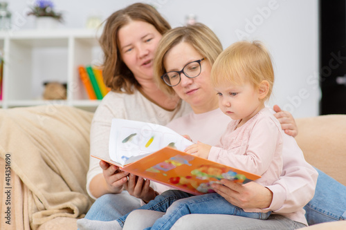 A homosexual family is reading a book to their child. Two girls and a baby are sitting at home on the sofa