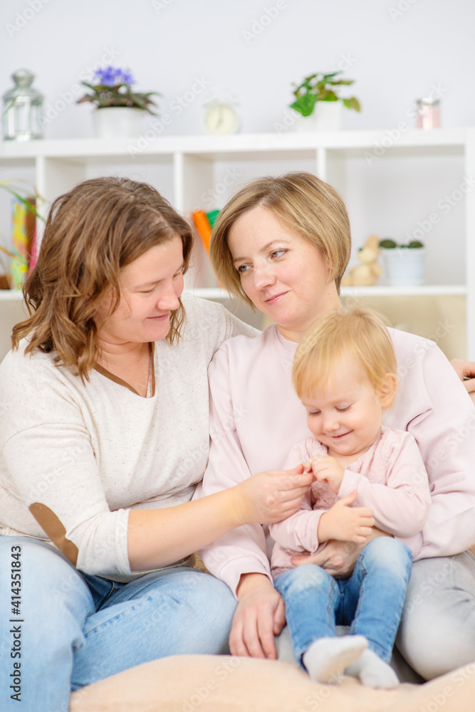 Two young women are sitting on the couch by the seyu at home in the living room and playing with their little daughter. Gay lesbian couple at home with a baby. Lesbian family Two young women are sitting on the couch by the seyu at home in the living room and playing with their little daughter. Gay lesbian couple at home with a baby. Lesbian family