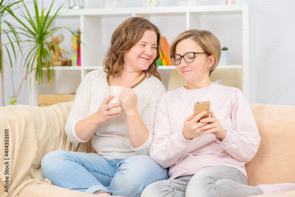 Two young women sit on the couch at home and look at the phone while having morning coffee. Gay lesbian couple at home. Lesbian couple concept
