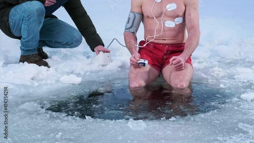 Medical test in the cold. Man sits on the ice with legs immersed into icy water. Oximeter, blood pressure monitor and cardio electrodes are connected to his body, another man comes and checks pressure photo