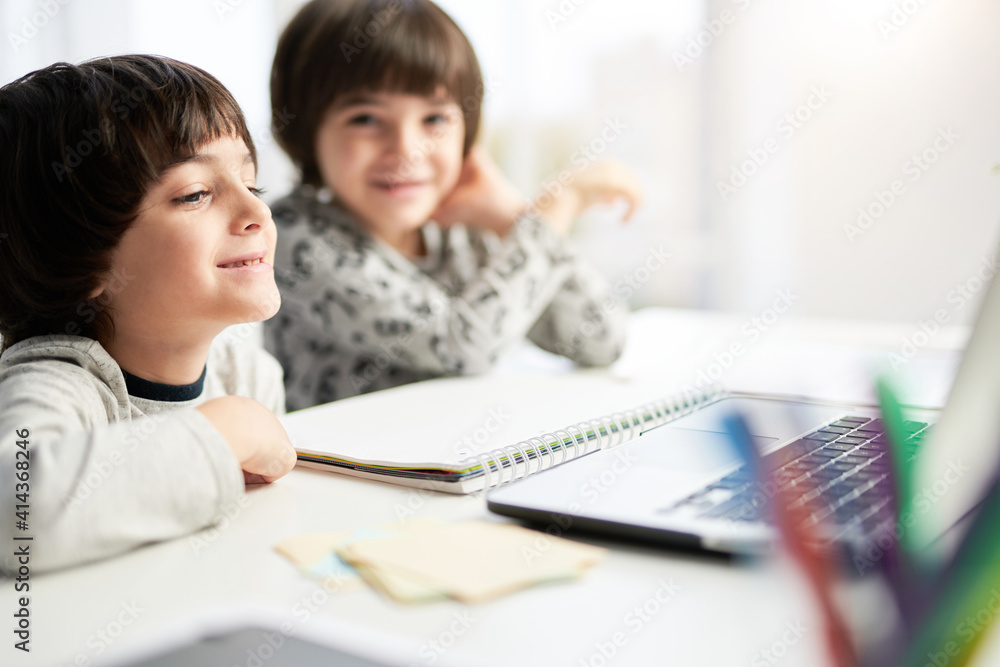 Learn at ease. Happy little hispanic boy looking at the laptop screen during online lesson for children, sitting with his twin brother at the table at home Learn at ease. Happy little hispanic boy looking at the laptop screen during online lesson for children, sitting with his twin brother at the table at home