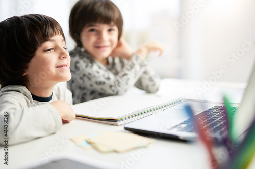 Learn at ease. Happy little hispanic boy looking at the laptop screen during online lesson for children, sitting with his twin brother at the table at home