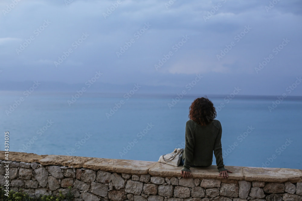 Lifestyle photo of young beautiful woman curly faceless looking at the calm blue sunset Mediterranean sea and dusk cloudy dramatic sky in Rhodes Island, Greece in summer. Soft focus, film edit. Lifestyle photo of young beautiful woman curly faceless looking at the calm blue sunset Mediterranean sea and dusk cloudy dramatic sky in Rhodes Island, Greece in summer. Soft focus, film edit.