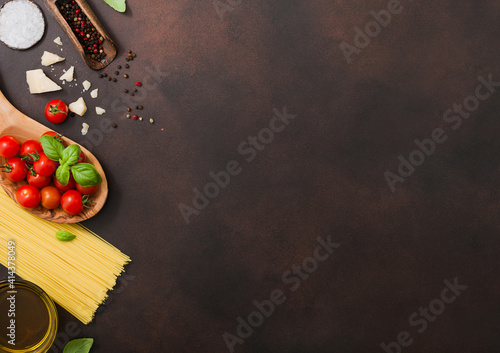 Spaghetti with cherry tomatoes in wooden bowl with pepper in scoop and parmesan cheese with salt on dark brown background.