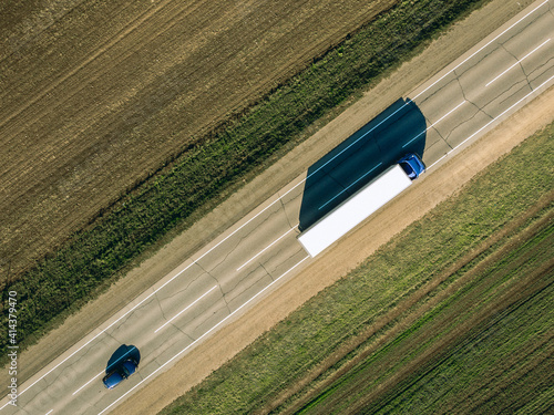 Bird's-eye view of a long paved road with trucks passing by. On the sides is cultivated land. Asphalt ground