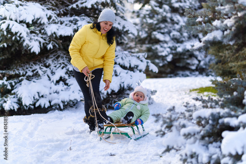mother and child walks in the winter forest, mom rides a baby on a sled, bright snowy fir trees, beautiful nature
