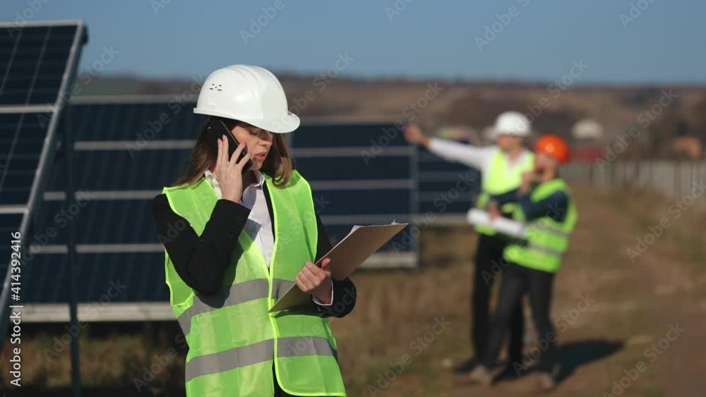A female engineer is talking on the phone. Two engineers are talking in the background. She is standing near the solar panels. 4K. A female engineer is talking on the phone. Two engineers are talking in the background. She is standing near the solar panels. 4K.