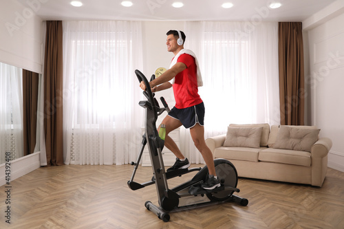 Man with headphones and towel using modern elliptical machine at home
