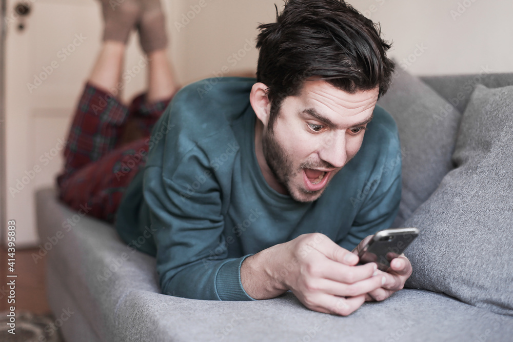 Young man holding a smartphone, looks very surprised and shocked, lying on a couch wearing pyjamas