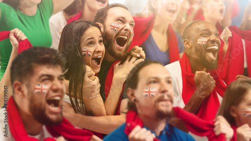 Britainian football, soccer fans cheering their team with a red scarfs at stadium. Excited fans cheering a goal, supporting favourite players. Concept of sport, human emotions, entertainment.