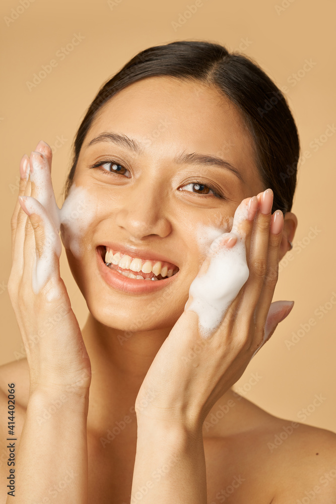 Beauty portrait of joyful young woman smiling at camera while applying gentle foam facial cleanser isolated over beige background Beauty portrait of joyful young woman smiling at camera while applying gentle foam facial cleanser isolated over beige background