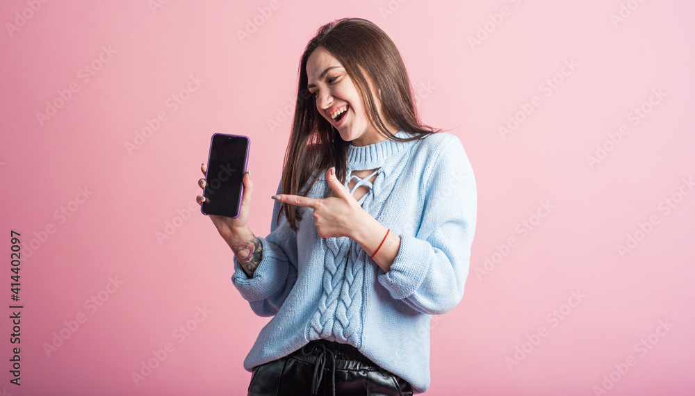 Brunette girl points a finger at a smartphone in the studio on a pink background Brunette girl points a finger at a smartphone in the studio on a pink background