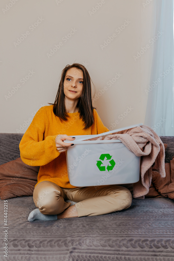 Woman sorting old clothes into white container with recycle sign. Green recycling sign on white container. Woman throwing out old clothes.