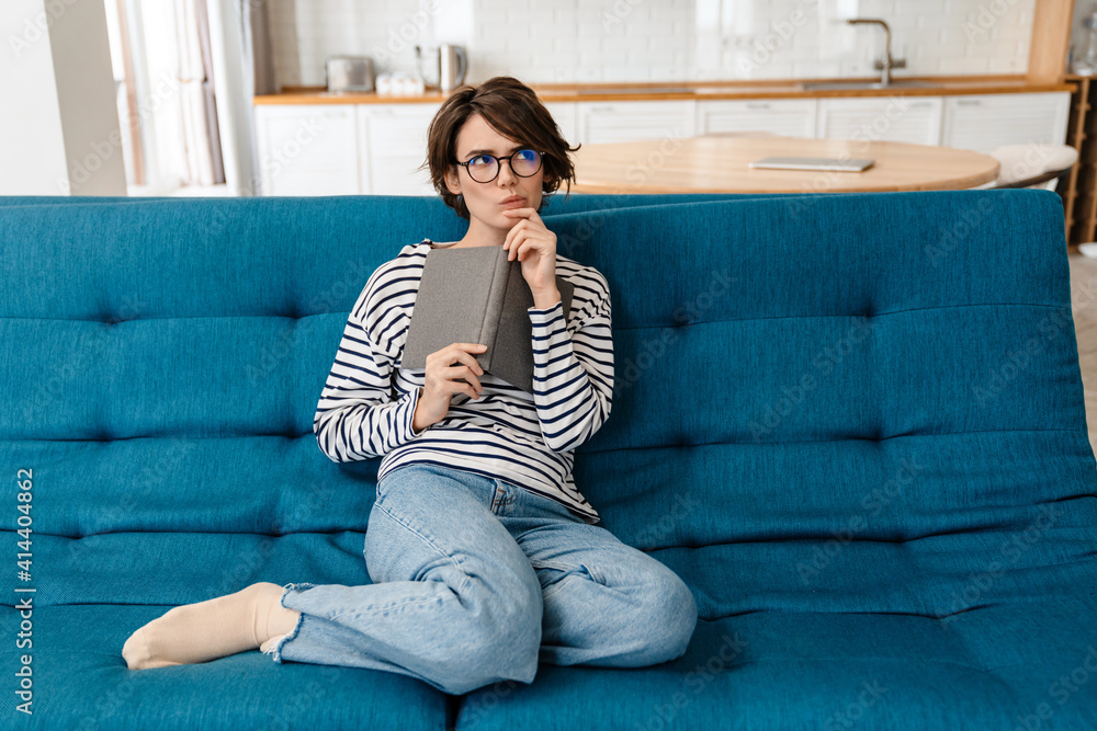 Brooding nice woman in eyeglasses reading book while sitting on couch Brooding nice woman in eyeglasses reading book while sitting on couch