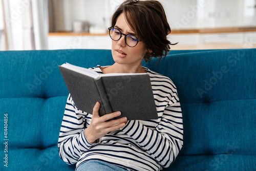 Brooding nice woman in eyeglasses reading book while sitting on couch