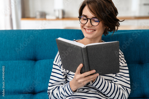 Happy beautiful woman in eyeglasses reading book while sitting on couch