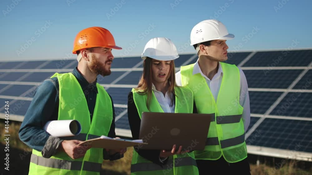 Three engineers are checking the data with a laptop and discussing it. They are standing next to solar panels. 4K. Three engineers are checking the data with a laptop and discussing it. They are standing next to solar panels. 4K.
