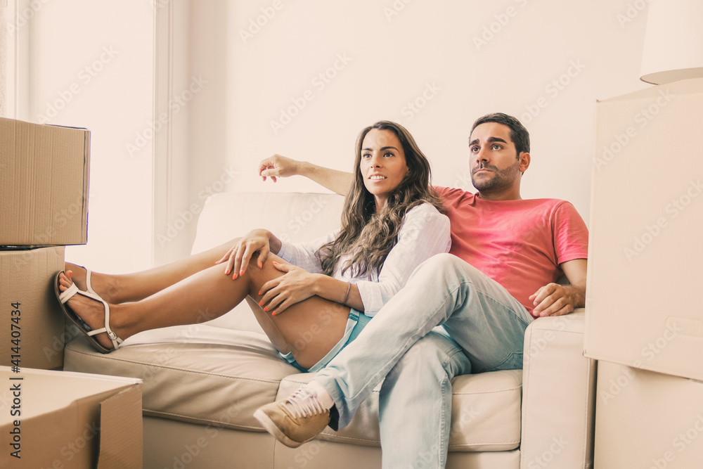 Positive Latin family couple relaxing on couch among carton boxes in apartment, looking away. Moving or new home concept Positive Latin family couple relaxing on couch among carton boxes in apartment, looking away. Moving or new home concept