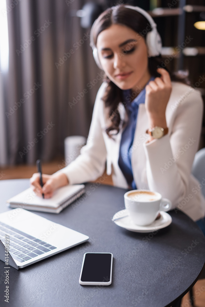 freelancer in headphones writing in notebook near laptop and smartphone with blank screen, blurred background freelancer in headphones writing in notebook near laptop and smartphone with blank screen, blurred background