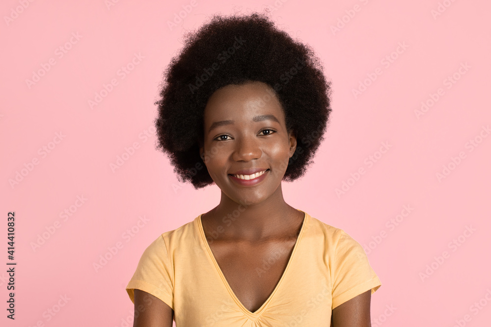 Portrait of young attractive african american woman, wearing yellow t-shirt, with afro hair and wide perfect smile on her face, posing to camera on pink pastel background. People, races concept