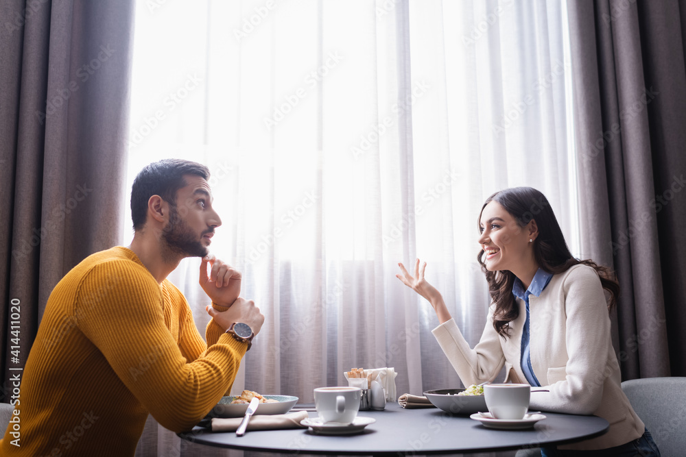 Smiling woman talking to arabian boyfriend near food and coffee in restaurant