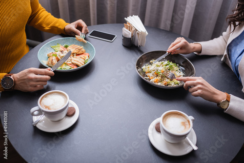 Cropped view of couple dinning with salads near cappuccino on blurred foreground in restaurant