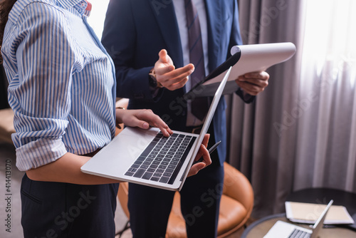 Cropped view of businesswoman using laptop near partner with clipboard on blurred background
