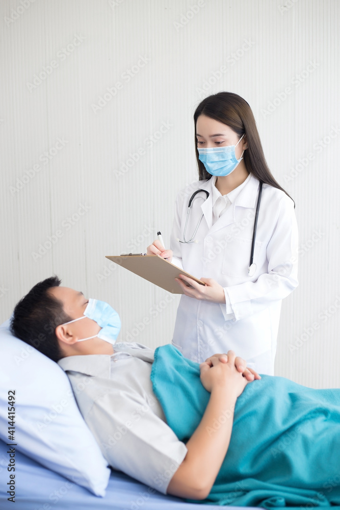 Asian woman doctor is diagnosing and recording health of her patient who an asian man and stay on bed in hospital. Both of them wear the medical face mask to protect Covid 19 virus. Asian woman doctor is diagnosing and recording health of her patient who an asian man and stay on bed in hospital. Both of them wear the medical face mask to protect Covid 19 virus.