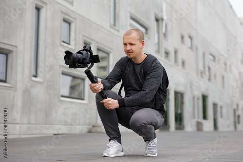 videography, filmmaking and creativity concept - professional male videographer shooting video using modern dslr camera on 3-axis gimbal over grey concrete building photo