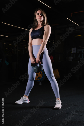 Young woman with athletic body poses standing in the gym with kettlebell.