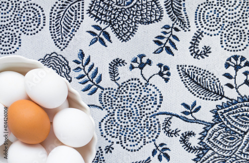 Many white eggs and one brown egg in a white dish against the background of a patterned tablecloth. Easter. top view